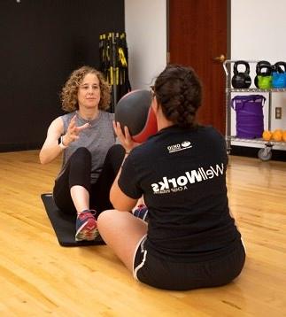 Two members passing a medicine ball between each other in a sitting position with their shoe tips touching and knees against their chest.